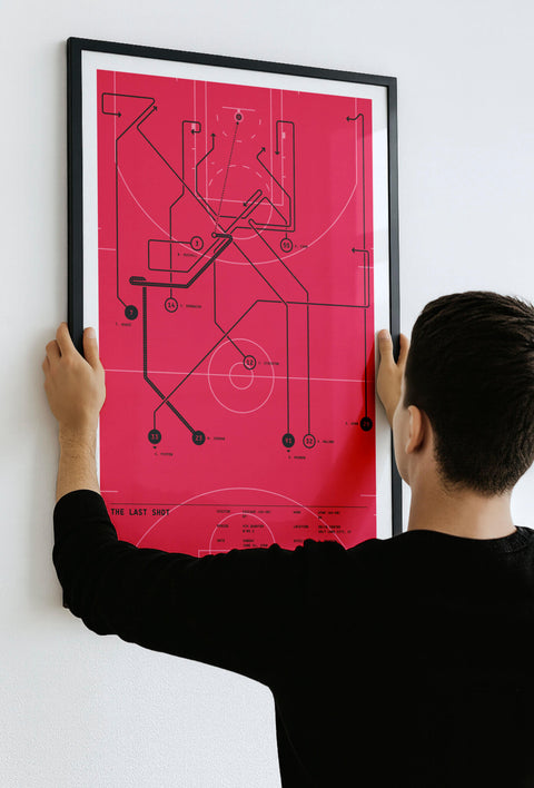 Man hanging a black-framed “The Last Shot” play diagram (red) on a white wall, showing ready-to-hang presentation.